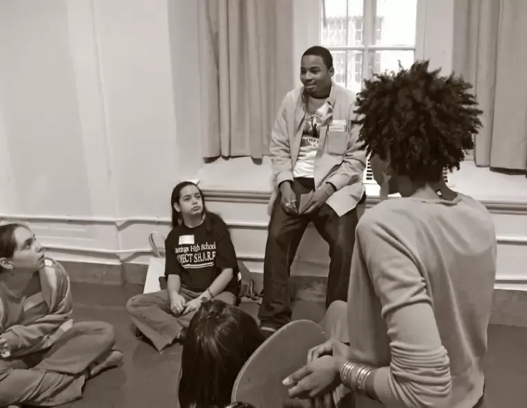 People in a circle, some seated on the floor, listening to a person with an afro. Indoor setting.