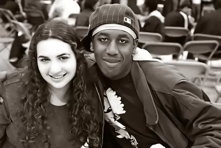 Two smiling people, a man with a cap and a woman with curly hair, pose close together in a room.