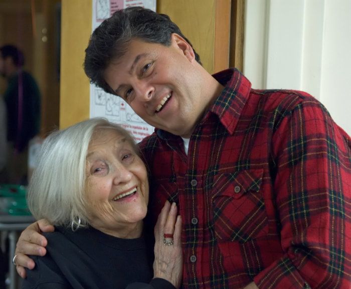 Man in red plaid shirt hugs smiling older woman indoors.