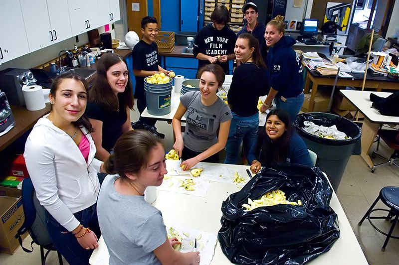 Group of people in a lab, peeling apples at a table. Trash bag and bucket nearby.