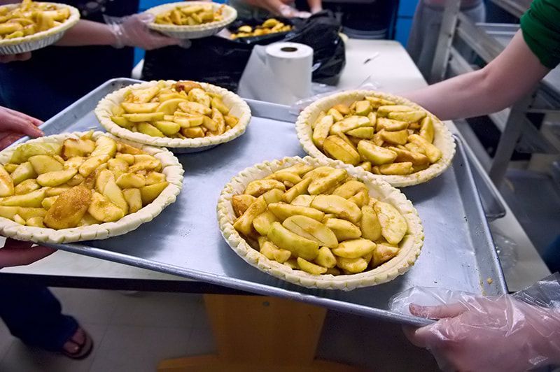 Apple pies in crusts on a tray, being held by people in a kitchen.