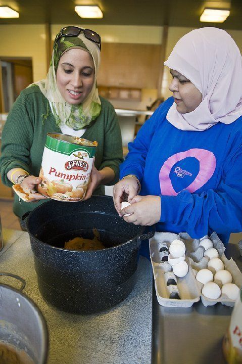 Two women wearing headscarves, preparing food together. One holds a pumpkin puree can, the other cracks eggs into a pot.