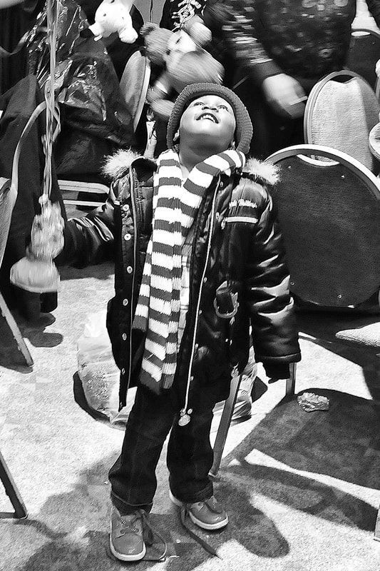 Child wearing scarf and jacket looks upward, surrounded by objects in an outdoor setting.