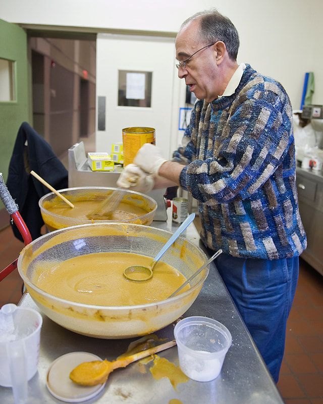 Man stirring a large bowl of orange liquid in a commercial kitchen; containers and utensils on a stainless steel counter.