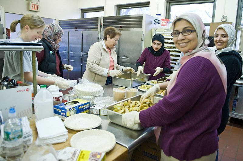 Volunteers prepare food in a kitchen. Women wearing headscarves and others in gloves smiling, working at counters.