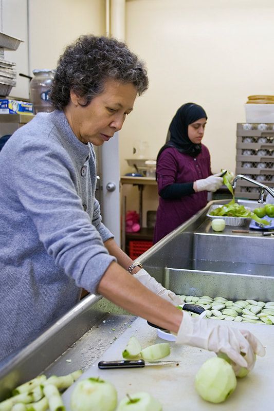 Two people peeling apples at a kitchen sink. One wears a gray sweater, the other a burgundy top and hijab.