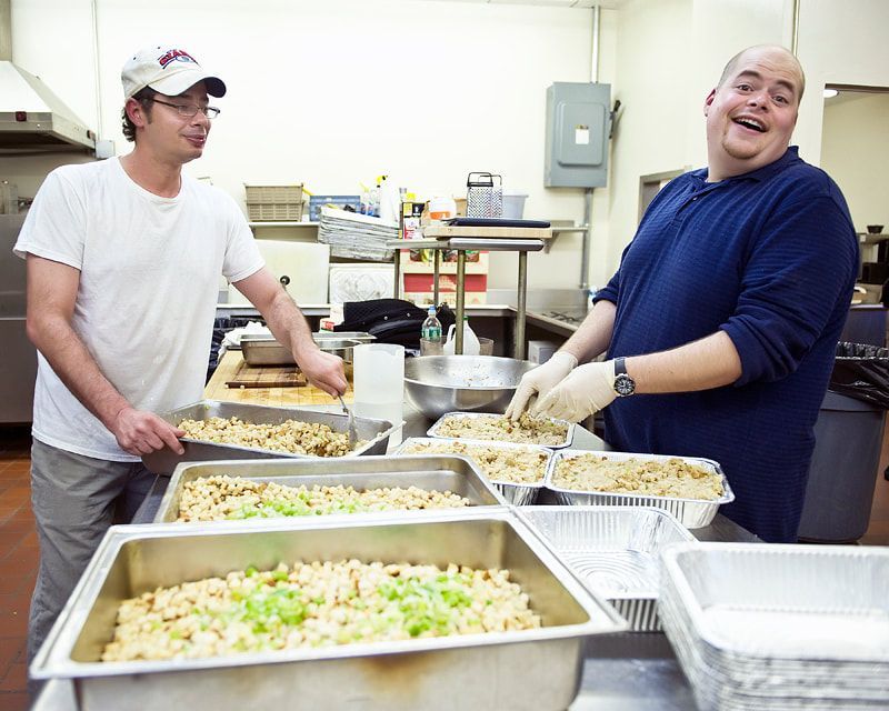Two men prepare food in a commercial kitchen, filling trays with stuffing. One smiles.