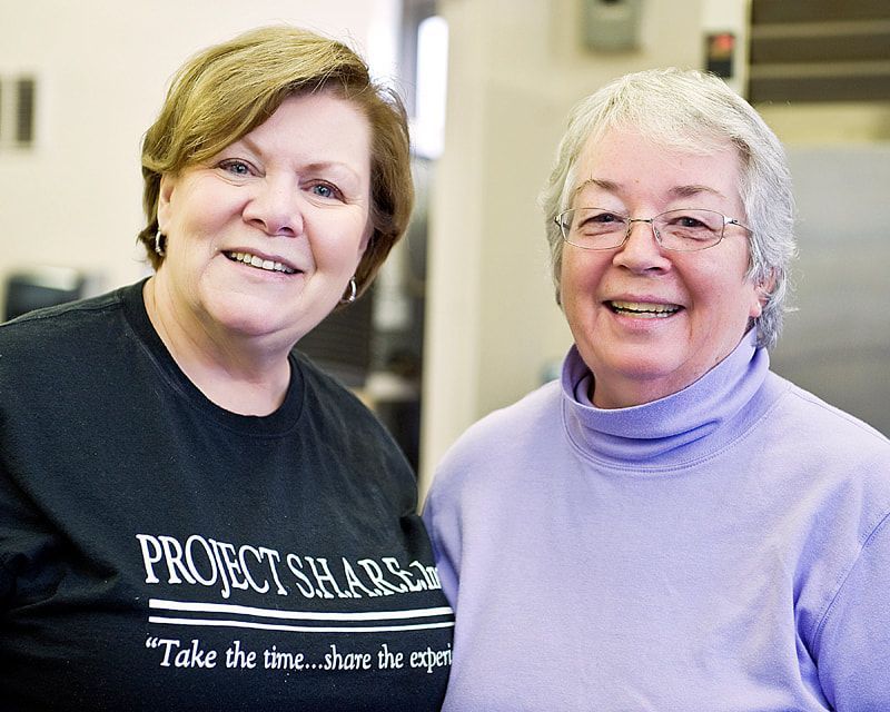 Two women smiling, one wearing a Project Share t-shirt, the other a purple turtleneck, indoors.