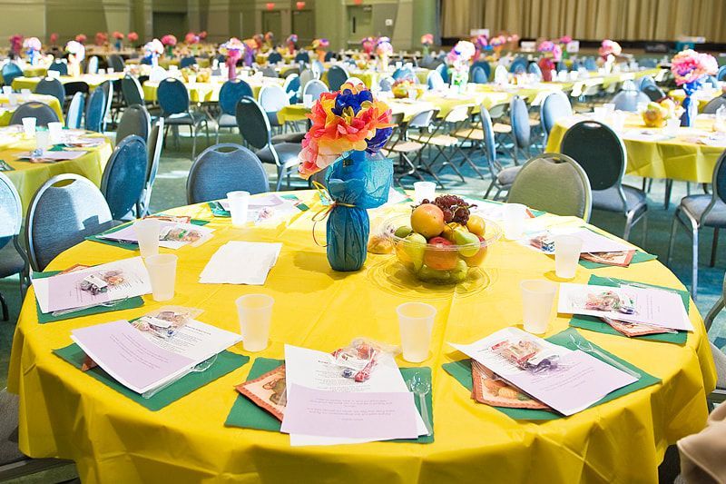 A banquet hall with yellow tablecloths, blue chairs, and colorful centerpieces, ready for guests.