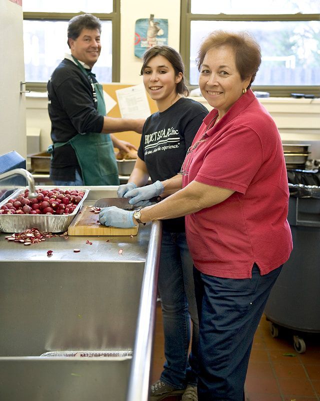 Three people preparing food in a kitchen. Two people wear gloves, while cutting produce. One person wears an apron.