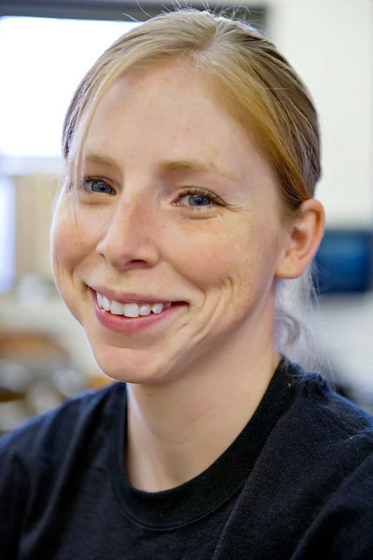 Woman with blonde hair, smiling, wearing a dark shirt, indoors.