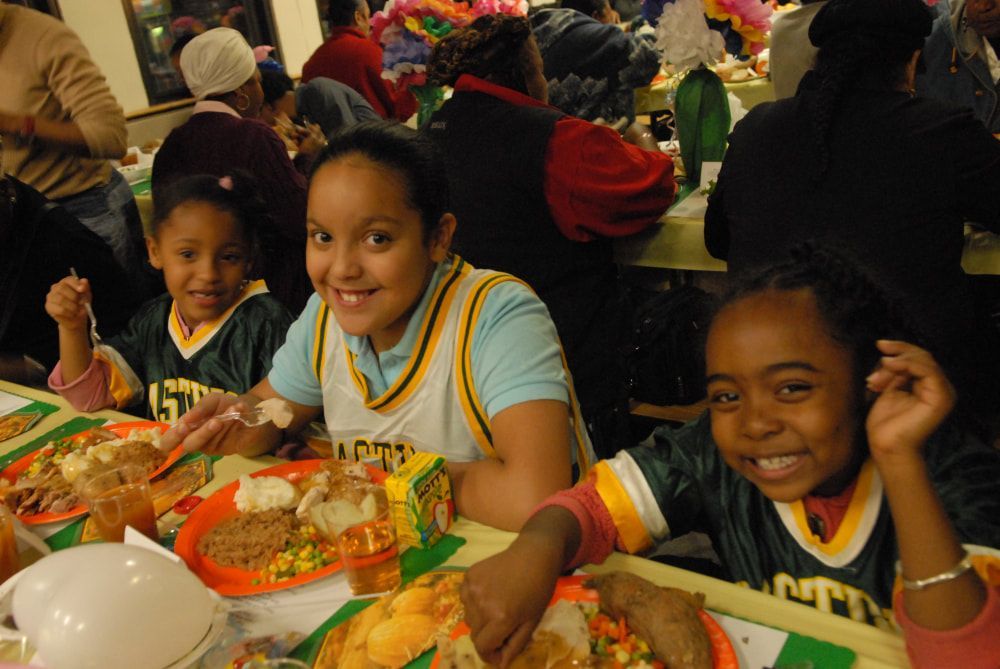 Three smiling children at a Thanksgiving dinner table with plates of food. Other people and decorations in the background.