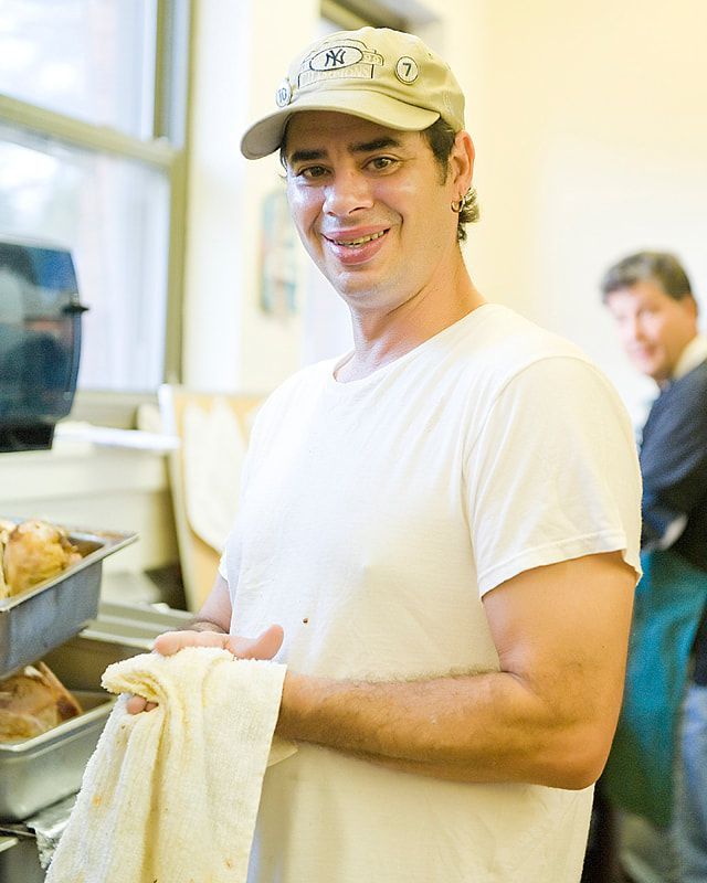 Man in baseball cap and white shirt smiles, holding a towel in a kitchen. Another man in the background.