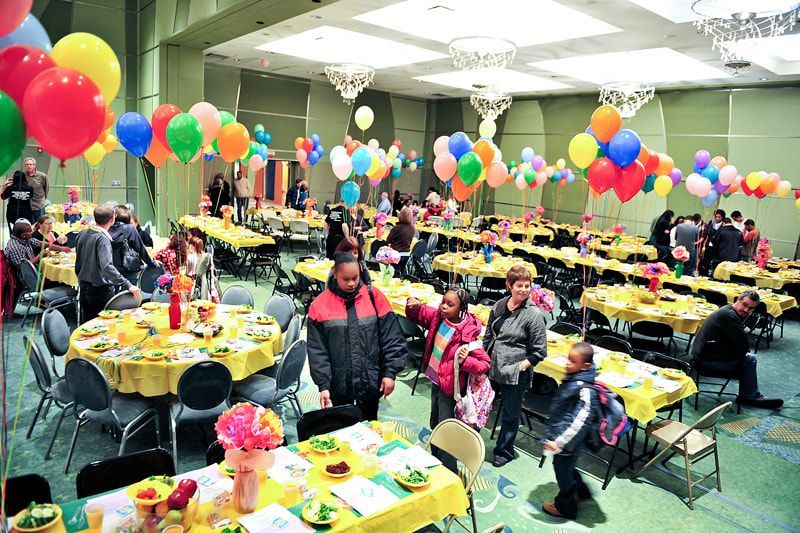 A banquet hall decorated with balloons and yellow tablecloths, filled with people standing and seated.