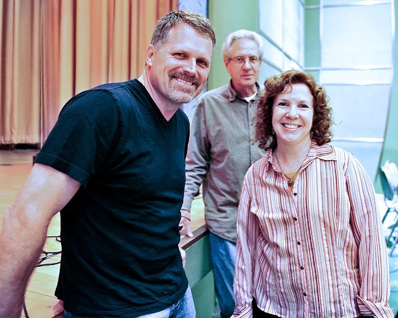 Three people smiling, inside a building. Man in black shirt, woman in striped shirt, older man behind.