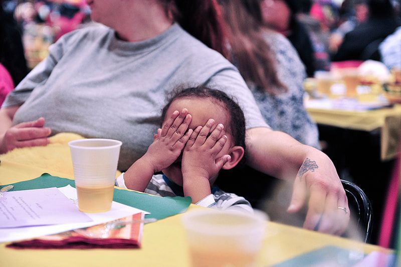 Child covering eyes, sitting at table with a woman. Drinks and papers on table.