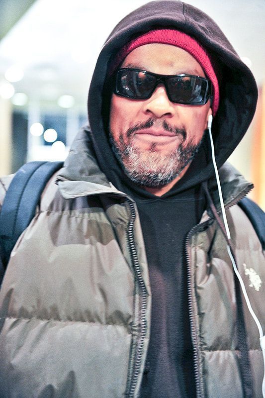 Man wearing sunglasses, red hat, and a puffer jacket, listening to music with earbuds.