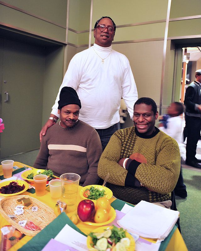 Three people at a table with food, drinks. One man stands behind them, smiling. Bright yellow tablecloth.