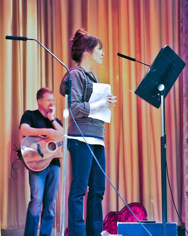 Woman with papers at a microphone on a stage, man with guitar in the background. Beige curtains.
