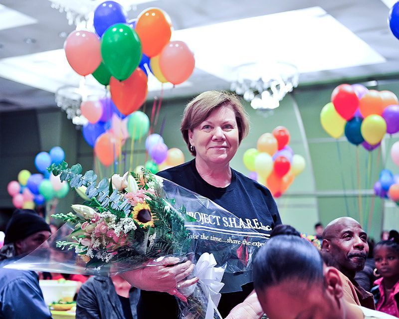Woman holding flowers smiles, surrounded by balloons and people at an event.