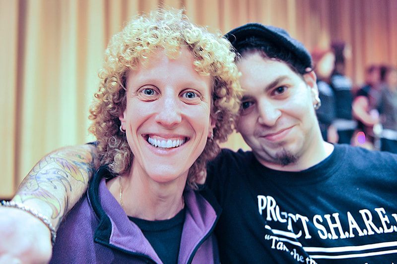 Two people smiling close together. One with curly hair, the other with a hat and goatee. Indoors.