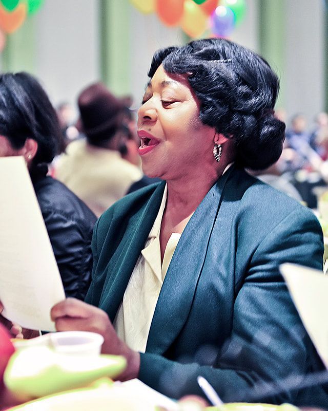 Woman singing, holding paper, wearing a green blazer, at a table with others, festive decorations.