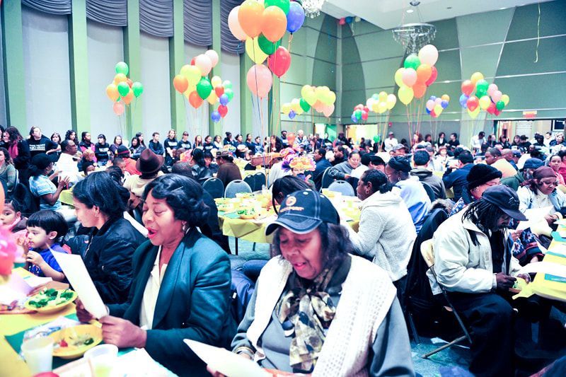 Large gathering in a decorated hall with tables, balloons, and many people seated.