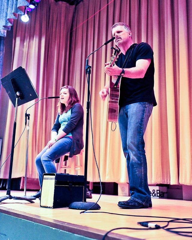 Man playing guitar, singing, and woman singing with him on stage; pink curtains.