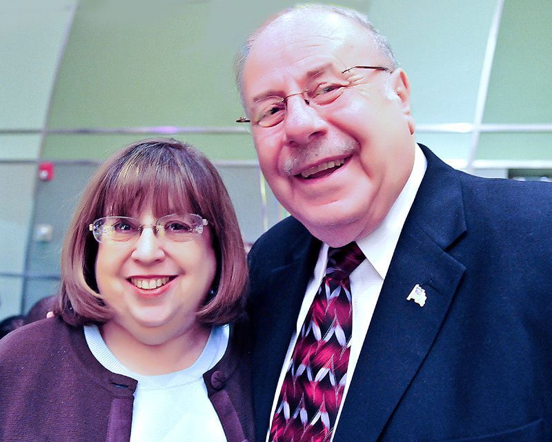 Woman and man smiling, close together. Woman wears glasses and brown sweater. Man in suit, tie. Indoors.