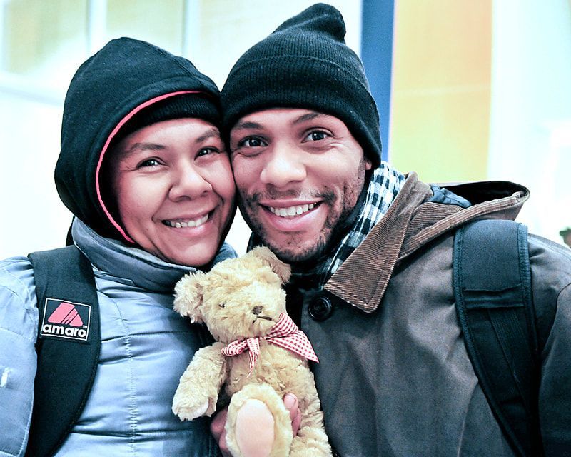 Smiling couple with a teddy bear, wearing winter hats and coats, with backpacks.