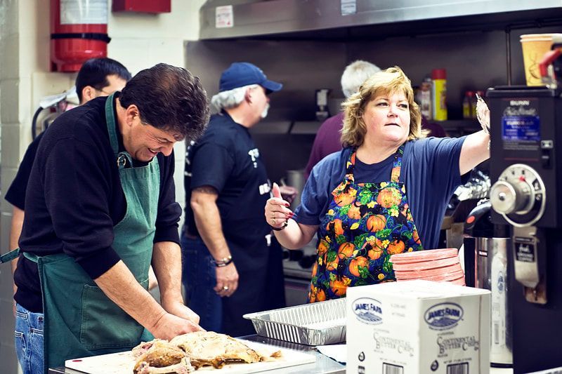 People in a kitchen preparing food. Woman gestures. Man carves meat. Others in the background. Stainless steel.