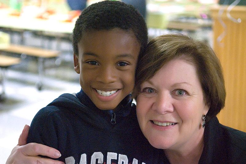 Boy smiles and hugs a woman; both look at the camera. They are indoors near tables.