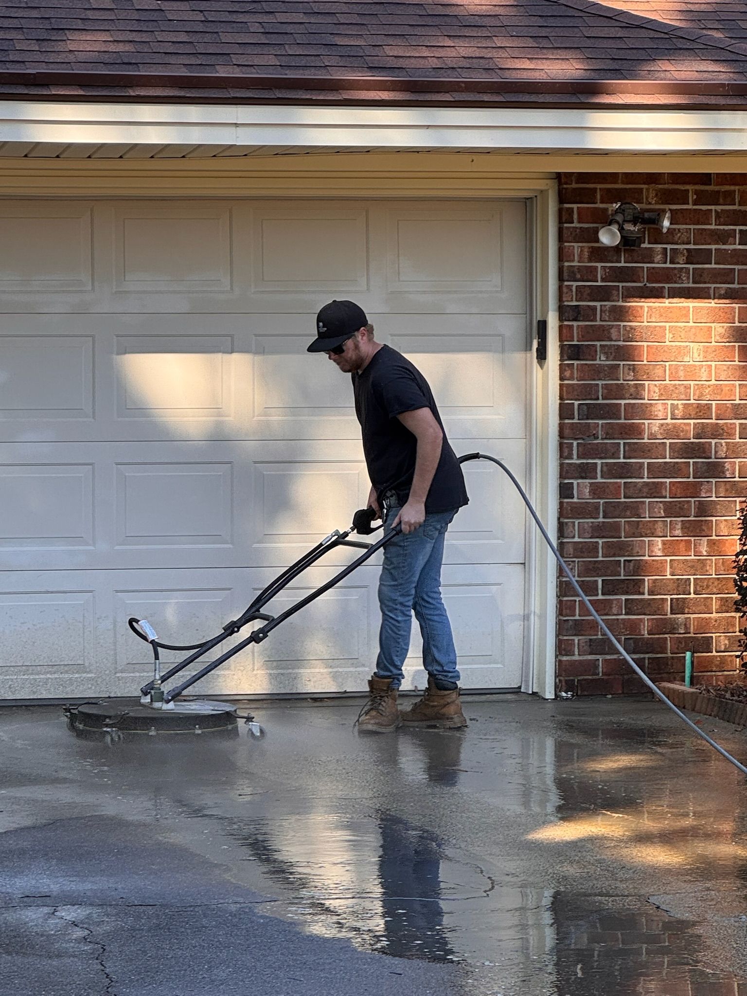 Man using a power washer on a wet driveway in front of a garage.