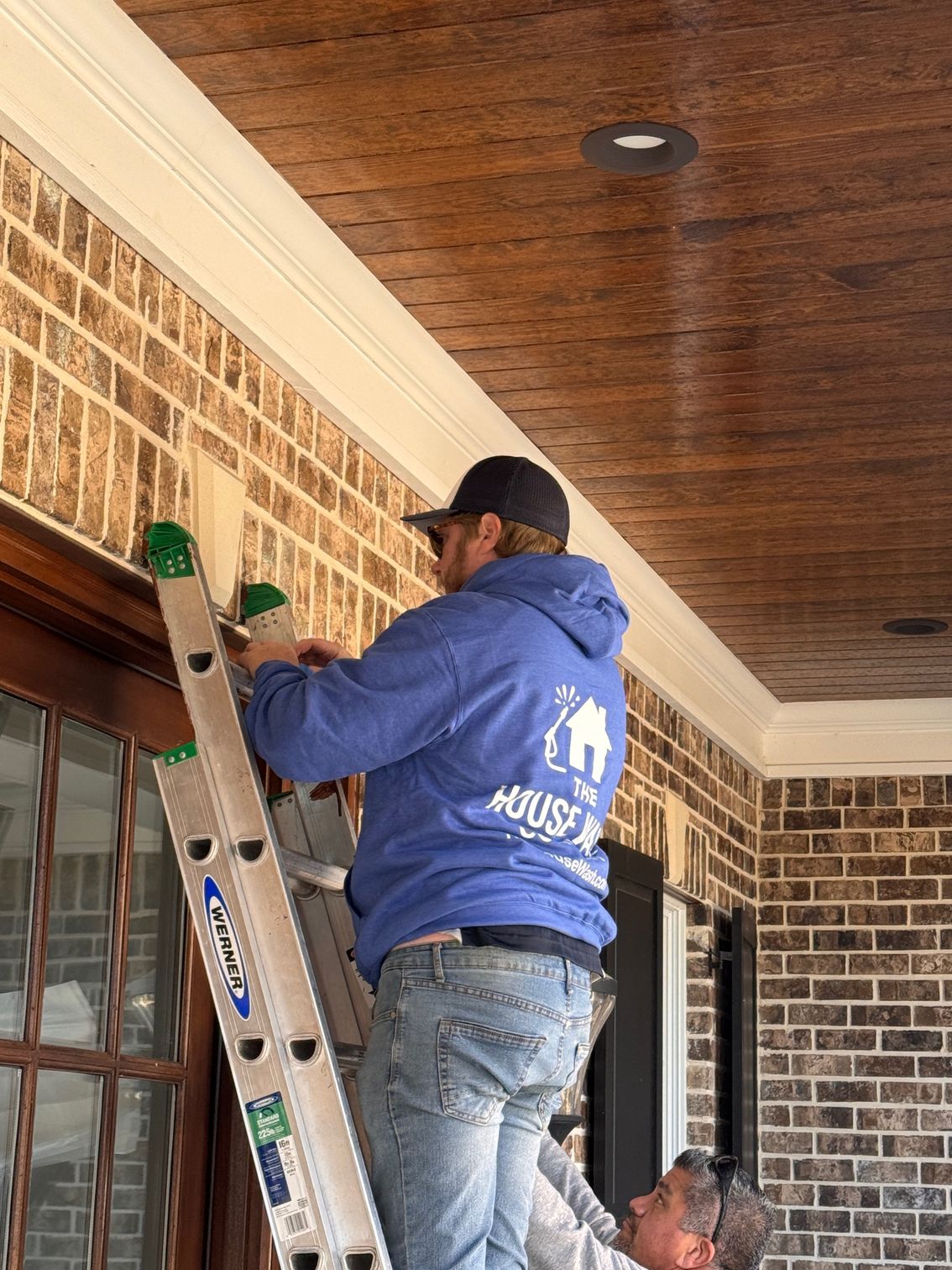 Man on ladder installing something on a brick building exterior. Another man helps below.
