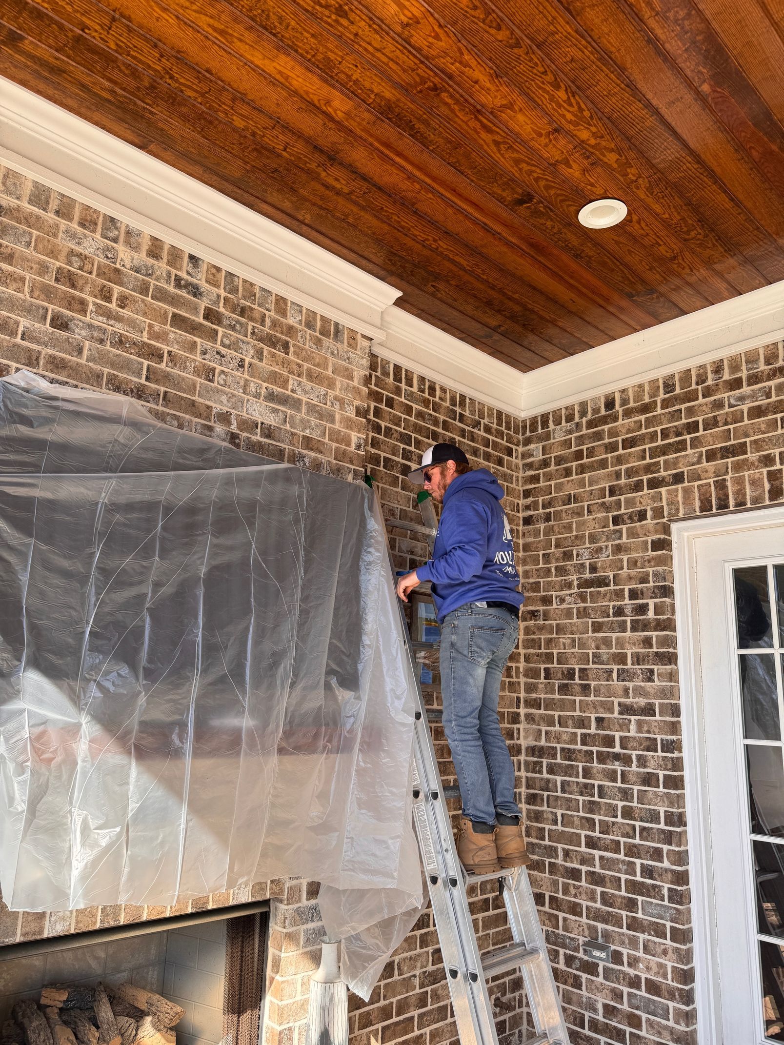 Man on ladder covering brick wall with plastic sheeting. Brown ceiling, white trim, and a doorway are visible.