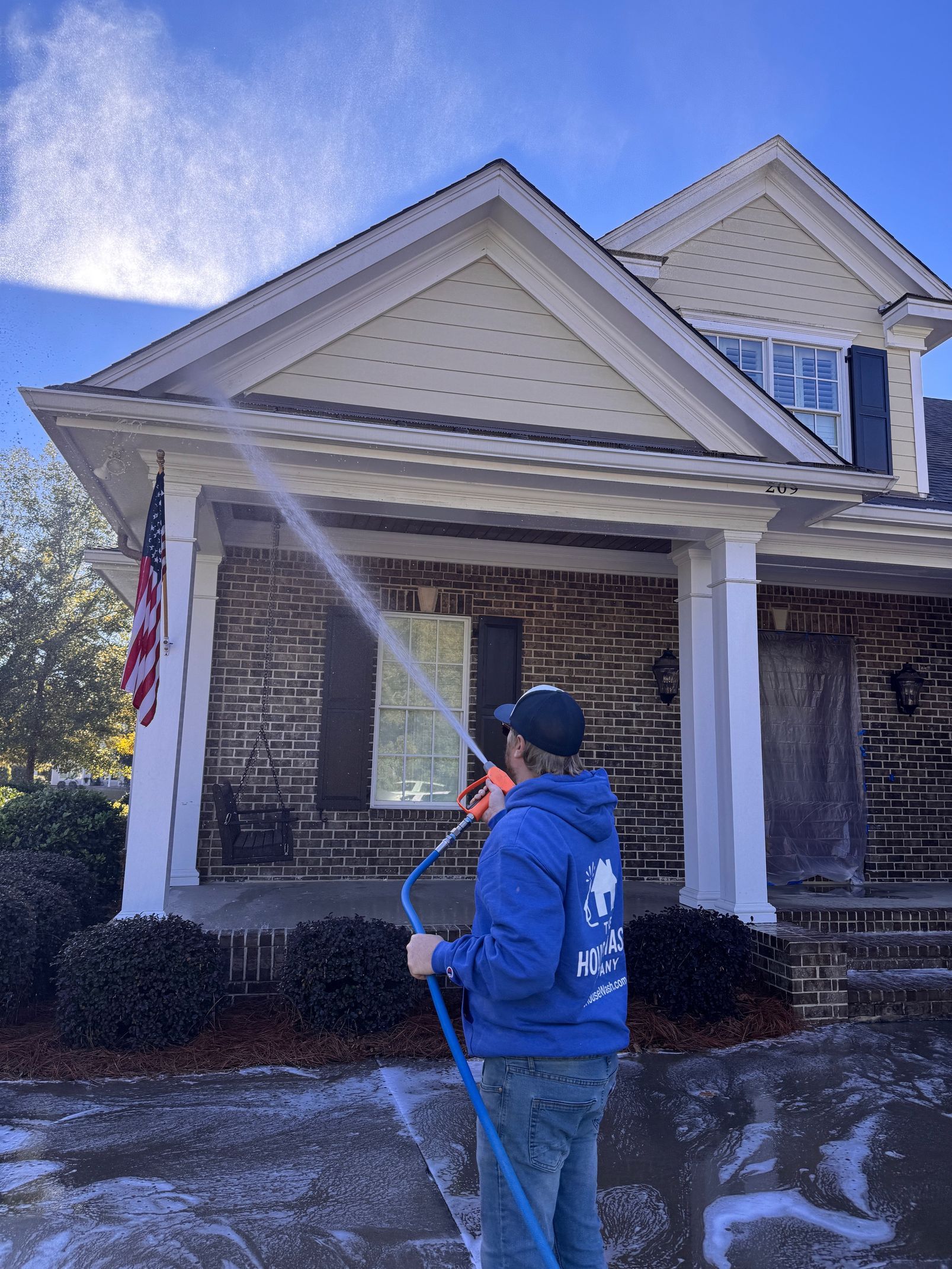 Person in blue jacket power washing a two-story house with a porch and American flag on a sunny day.