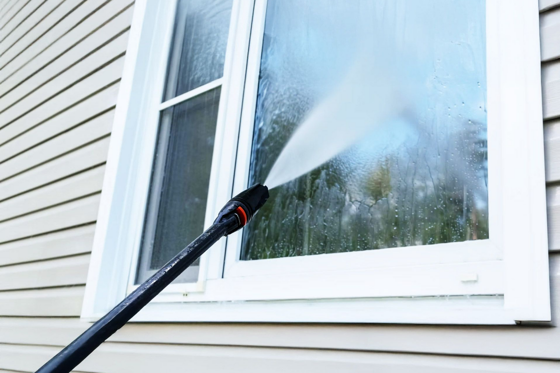 Pressure washing a window on a house with gray siding.