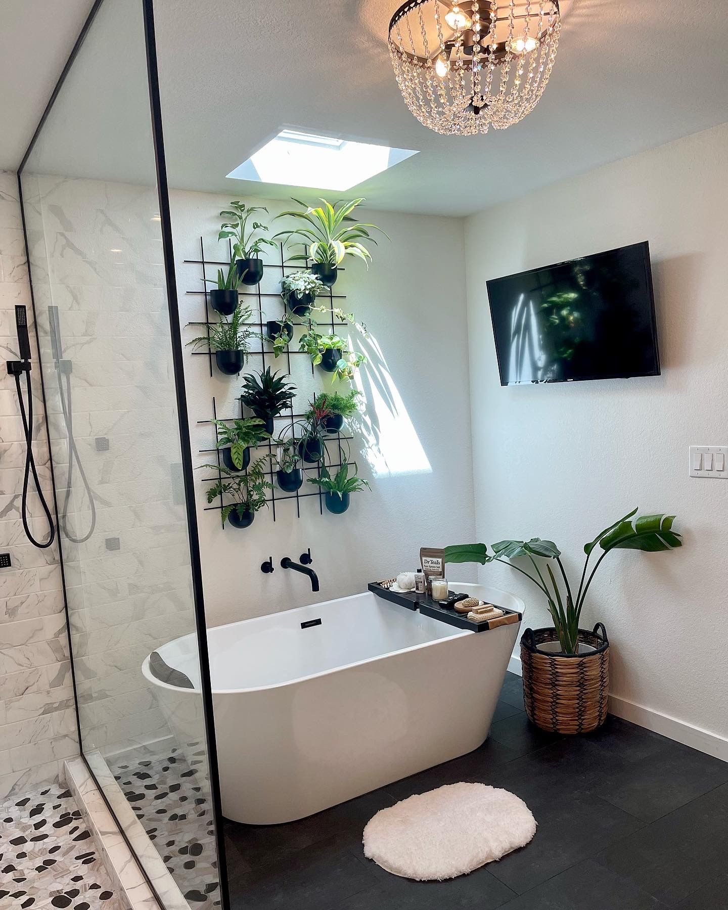 Modern bathroom with tub, shower, and wall of plants. A tv and chandelier light the space.