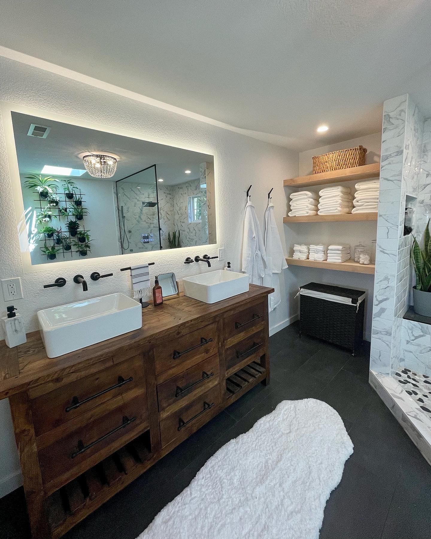 Modern bathroom with a wooden vanity, large mirror, marble accents, and shelves with towels and toiletries.