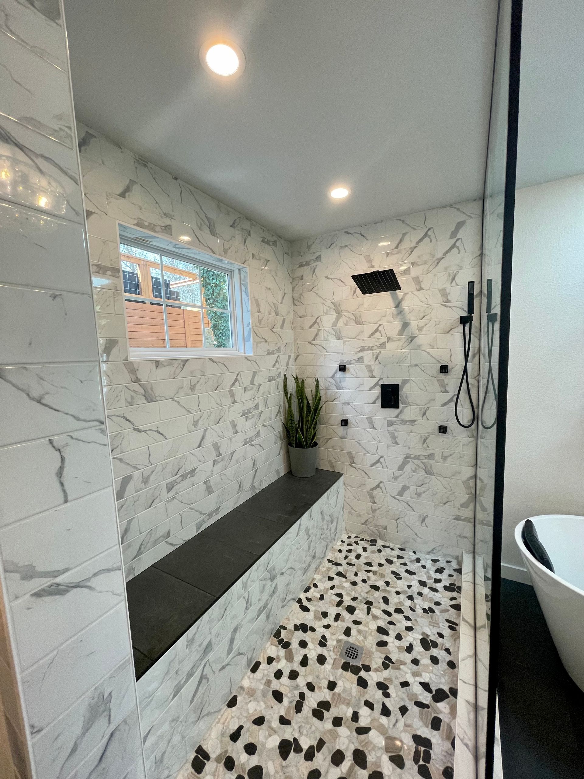 A modern shower with marble tile, pebble floor, built-in bench, glass enclosure, and black fixtures.