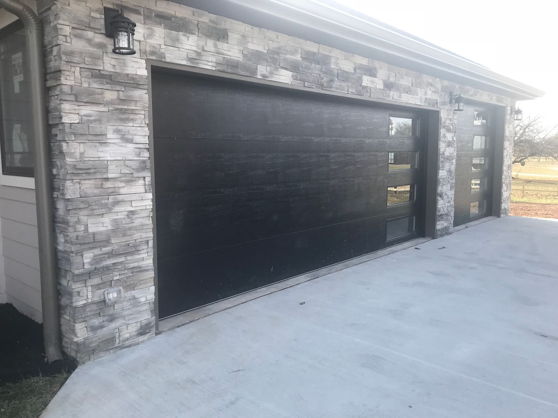 Black garage doors with modern glass windows, set in a gray stone facade with concrete driveway.