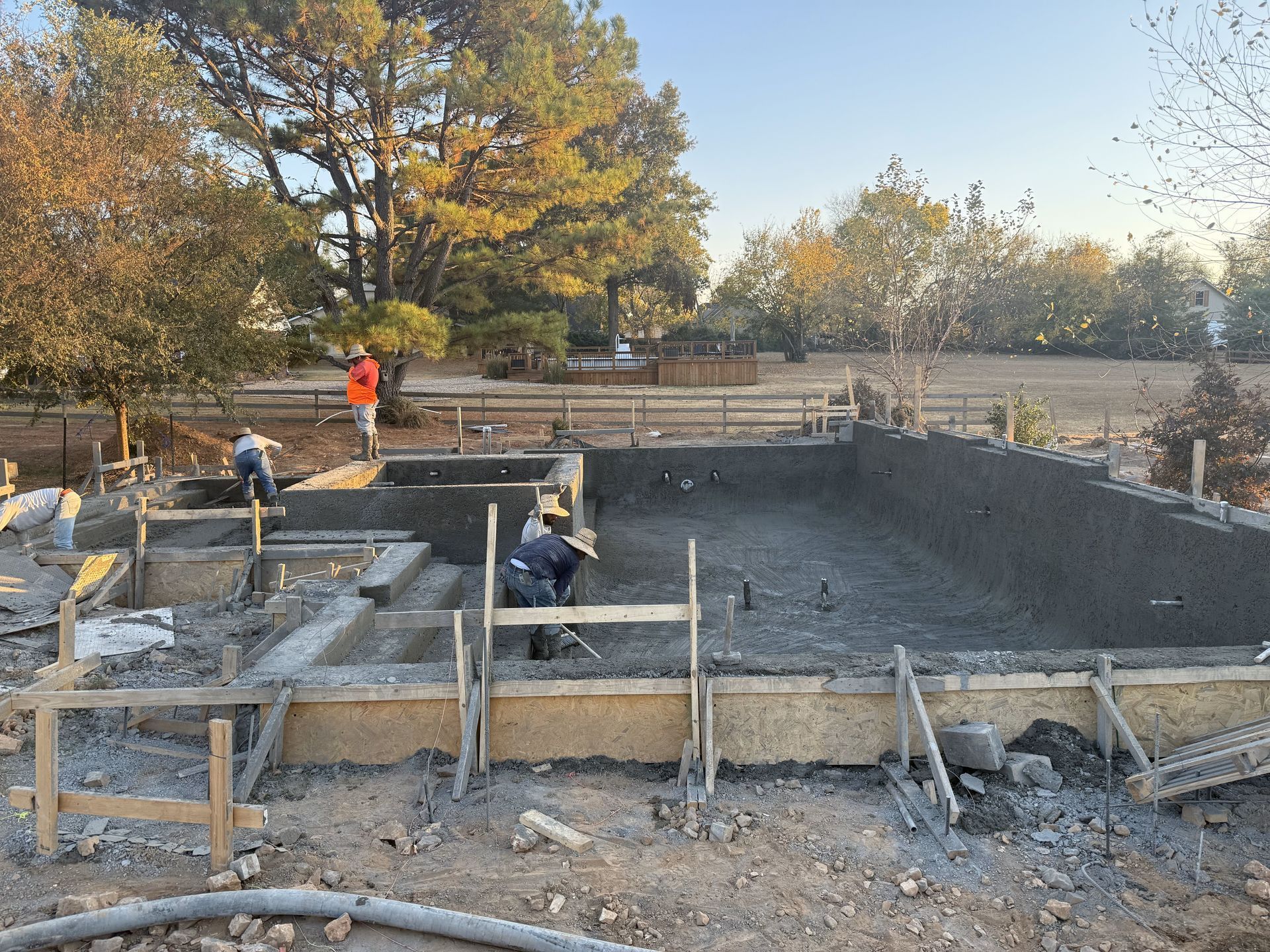 Construction workers pouring concrete into a rectangular pool, with surrounding forms and a rural setting.