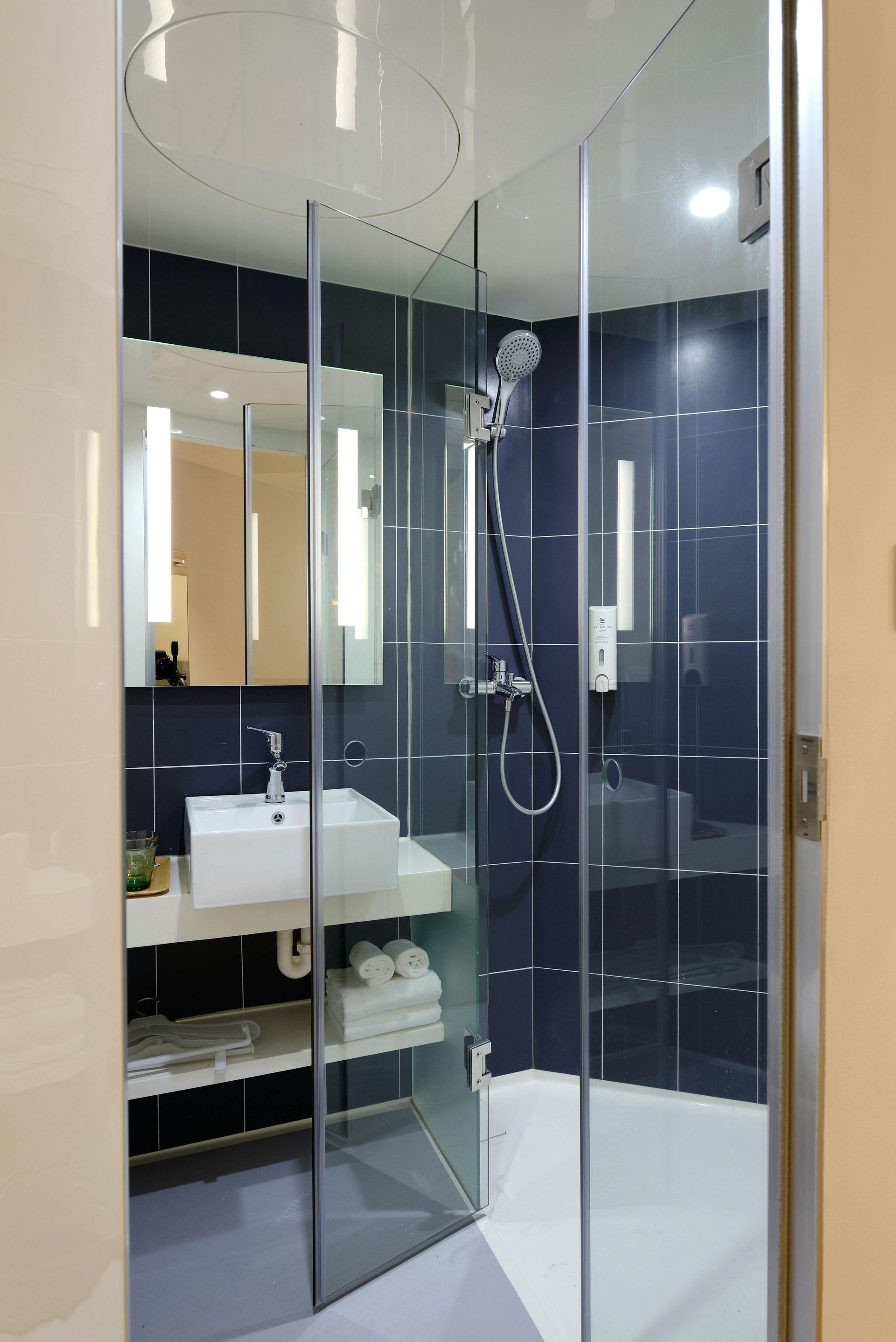 Small modern hotel bathroom with shower, sink, and mirror. Dark blue tile, white sink, glass shower door.