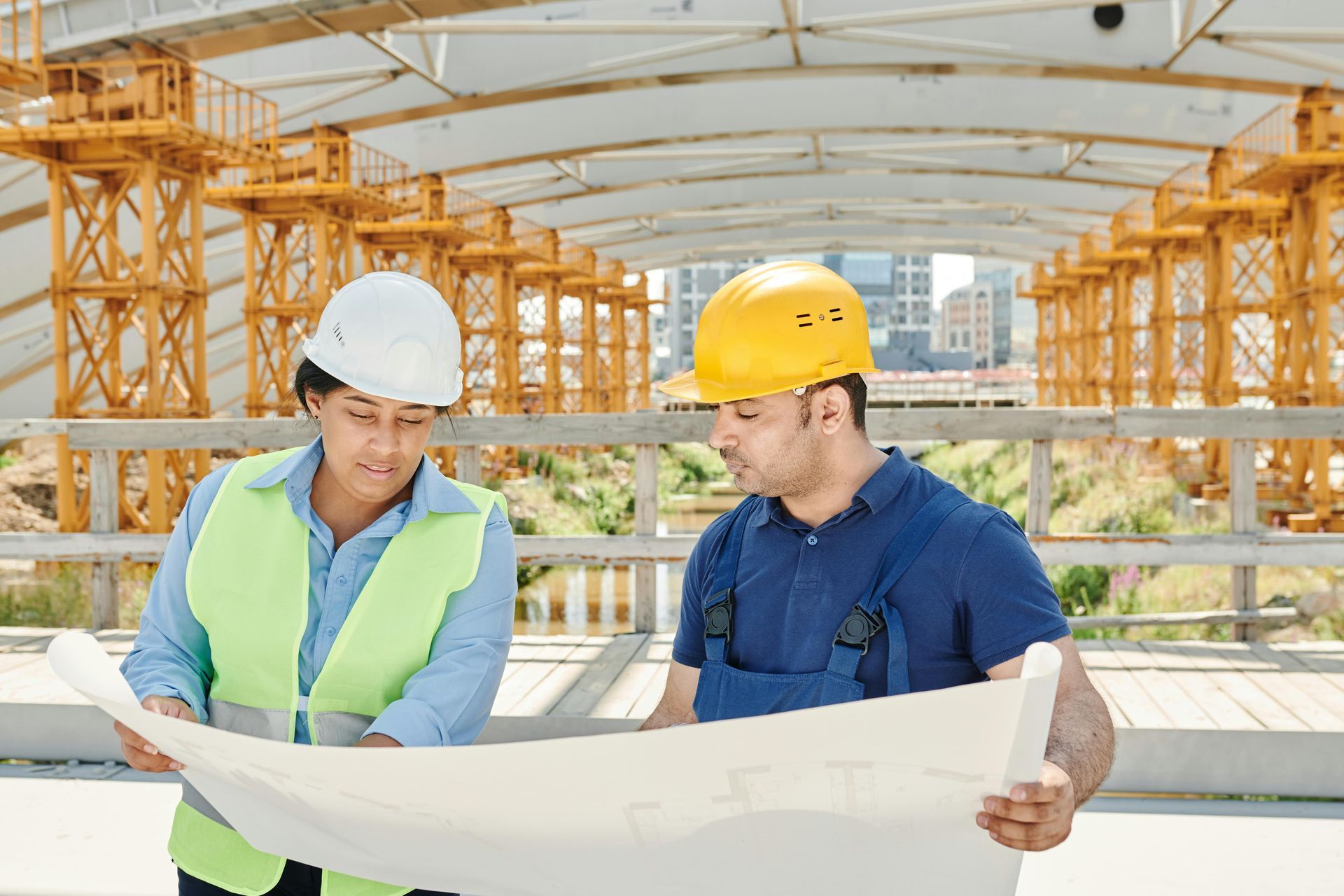 Two construction workers reviewing blueprints on a commercial construction site, both wearing hard hats and safety vests.