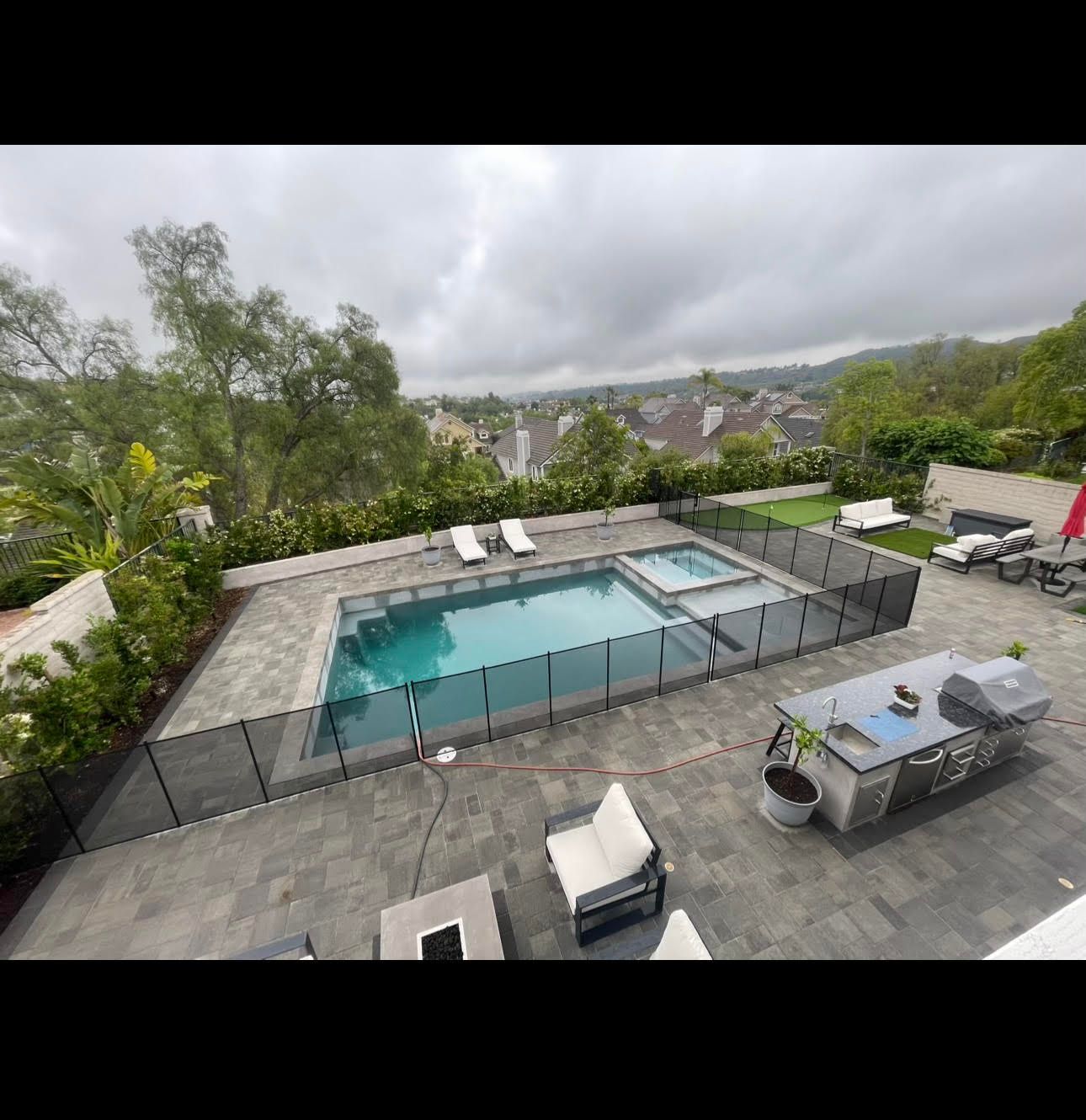 Swimming pool area with dark stone patio, black fence, and outdoor kitchen under cloudy skies.