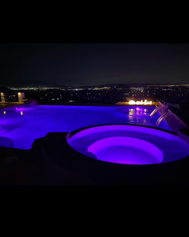 A pool and jacuzzi at night with purple lights overlooking a city skyline and fire.