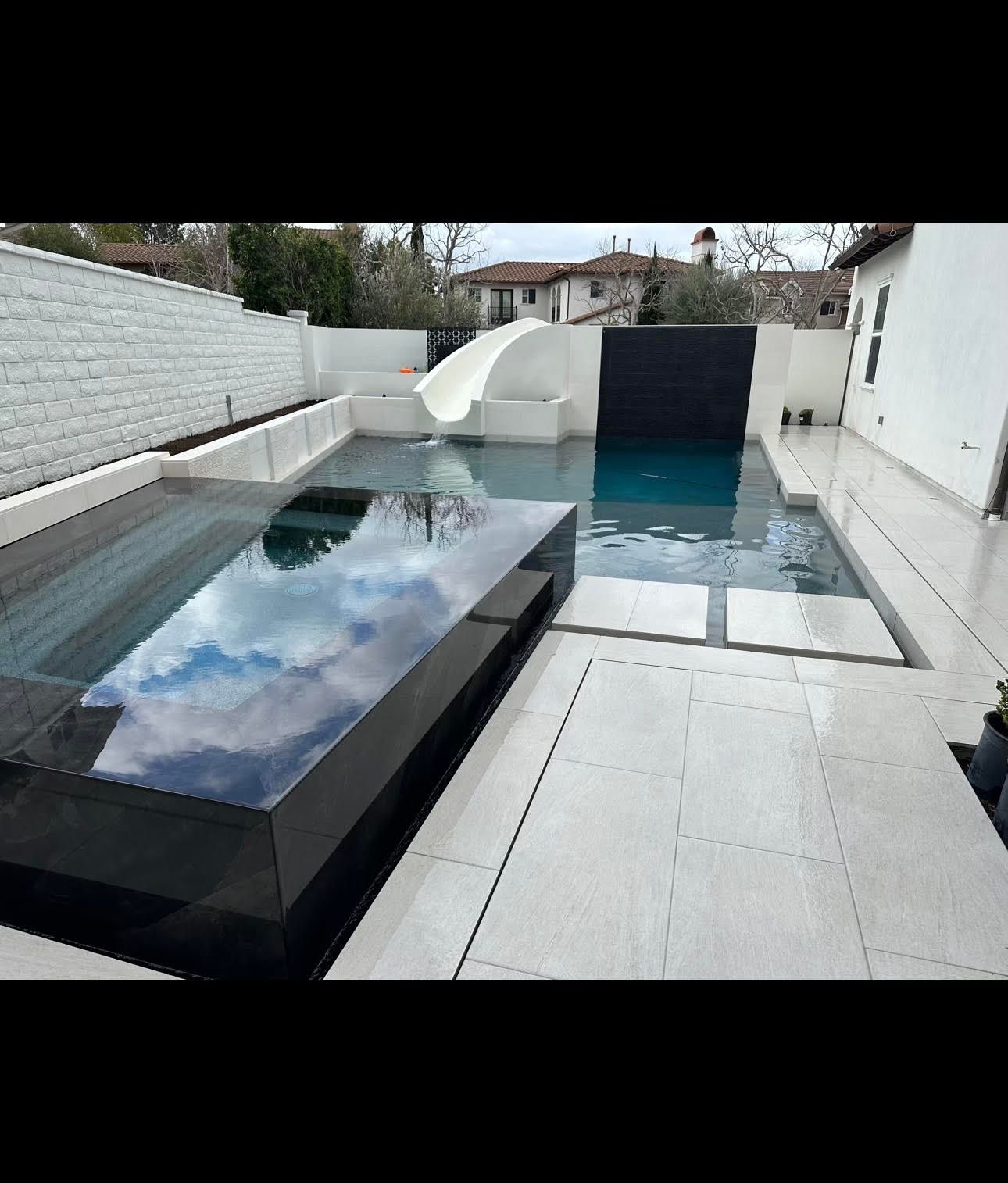 Modern backyard pool with water slide, dark jacuzzi, and white patio tiles. Cloudy sky reflections.