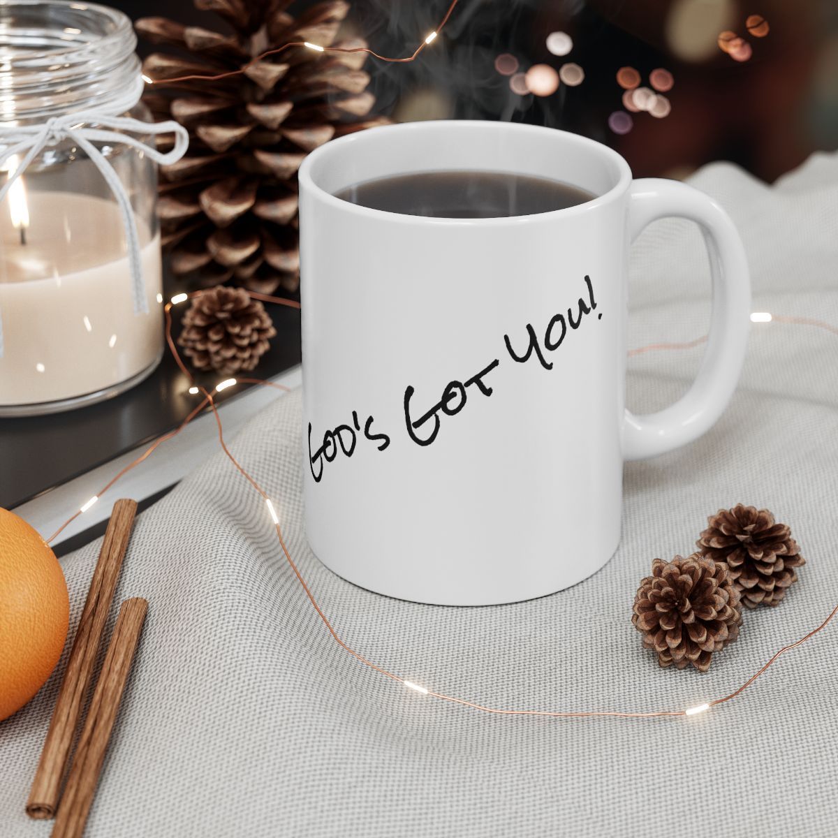 White mug with "God's for You!" text, coffee inside, beside candle, pine cones, orange, and cinnamon sticks on fabric.