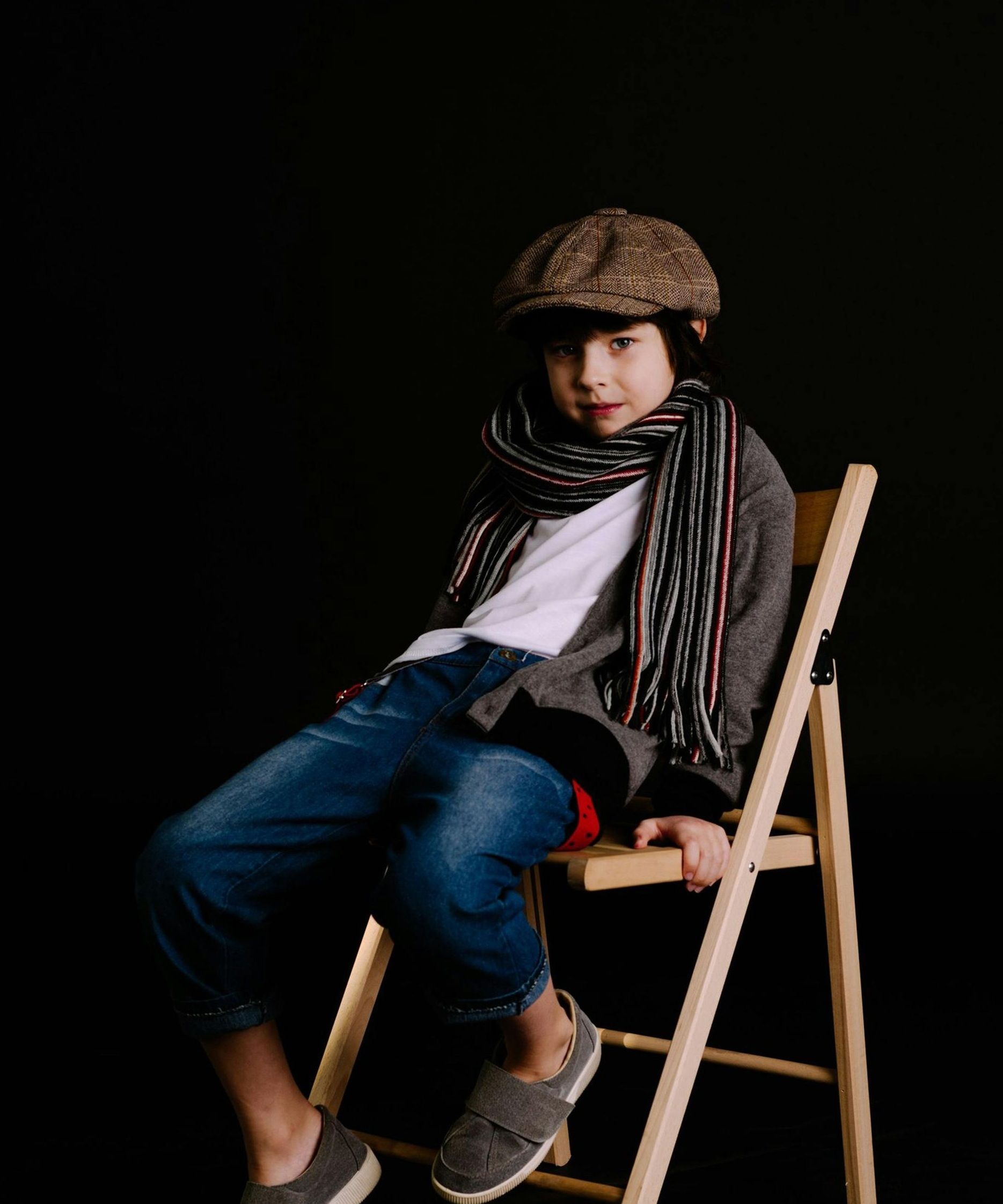 Young person in a hat and scarf sits on a wooden chair against a black background.