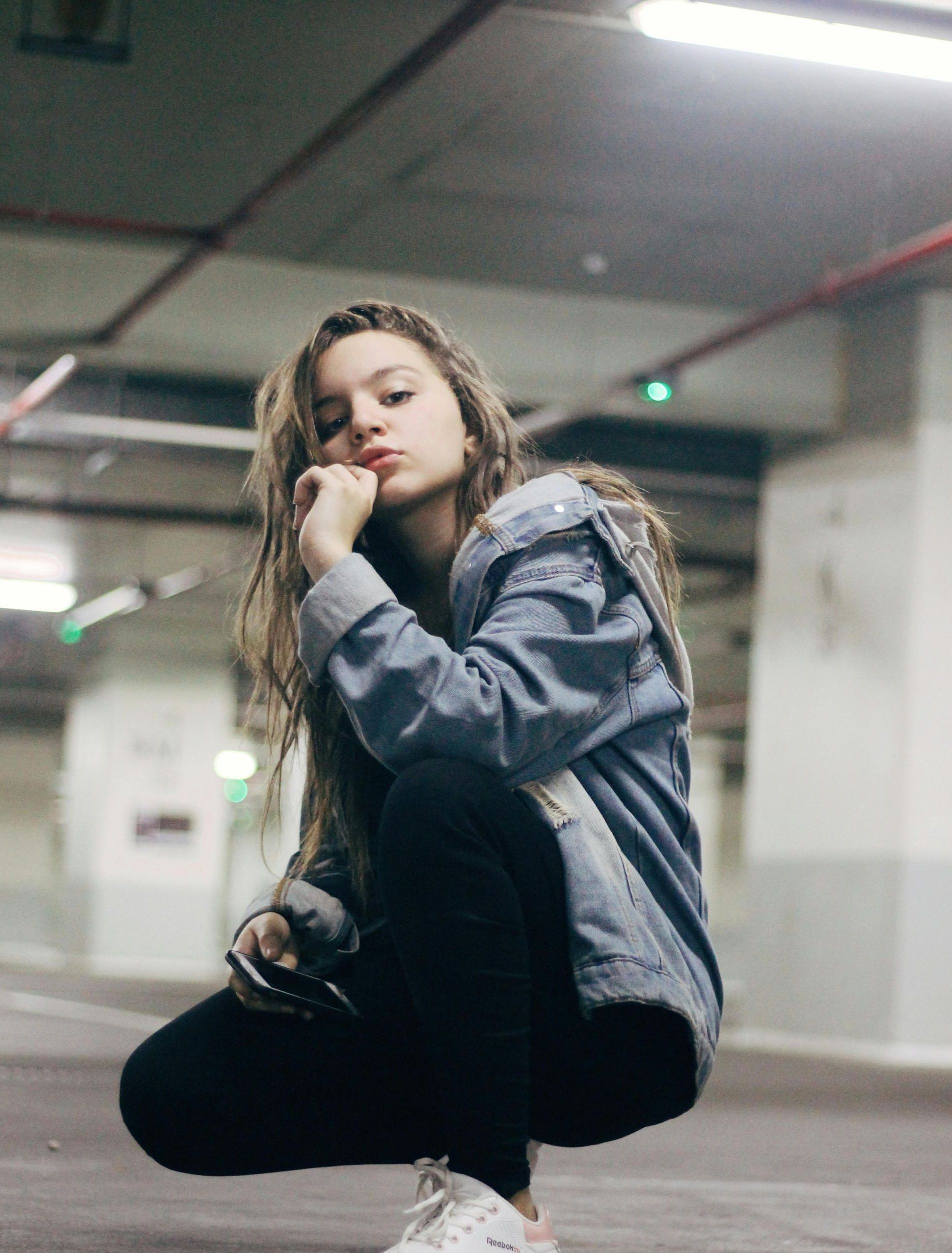 Young woman in a denim jacket and black pants squats in a parking garage, looking at the camera.